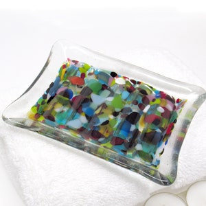 May include: A clear glass soap dish with a colorful mosaic design. The mosaic features various shades of blue, green, red, yellow, and black.