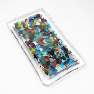 May include: A rectangular glass dish with a clear base and a colorful mosaic design. The mosaic features small pieces of glass in shades of blue, green, red, yellow, and black.