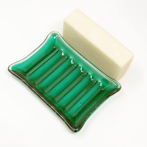 May include: A handmade, emerald green glass soap dish with a rectangular shape and raised ridges. A bar of unscented soap rests on the dish. The soap dish is a decorative and functional bathroom accessory.