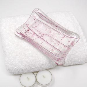 May include: A clear glass soap dish with pink accents rests on a white, fluffy towel. The dish has a rectangular shape with curved edges and pink lines and speckles. Two small, white candles are at the bottom.