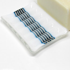 May include: A white glass soap dish with a blue and black striped design. The dish has a slightly curved shape and is designed to hold a bar of soap.
