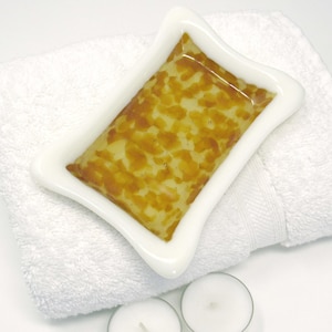 May include: A white ceramic soap dish with a rectangular, star-shaped design holds a bar of soap with a yellow and brown pattern. The dish rests on a folded white towel, with two small white candles in the foreground.