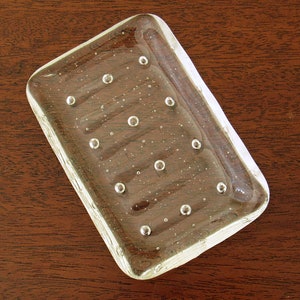 May include: A clear glass soap dish with a rectangular shape and a textured surface. The dish has small air bubbles throughout the glass.