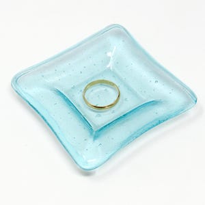 May include: A light blue square glass dish with a ring inside. The dish has a textured surface with small bubbles.