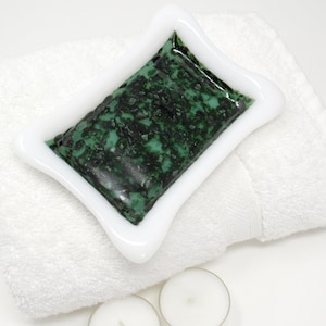May include: A white, star-shaped soap dish with a green and black soap bar, resting on a white towel. Two small, white candles are at the bottom of the image. The soap dish has a decorative, curved edge.
