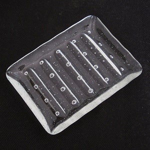 May include: A rectangular clear glass soap dish featuring a series of raised parallel ridges across the surface. The glass contains numerous small embedded air bubbles throughout the structure, creating a textured appearance across the entire piece.