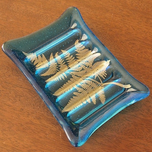 May include: A blue glass soap dish with a gold fern design. The dish has a rippled texture and is perfect for holding soap or other small items.