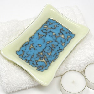 May include: A small, rectangular dish made of glass with a light yellow background and a blue and brown abstract design. The dish is sitting on a white towel.