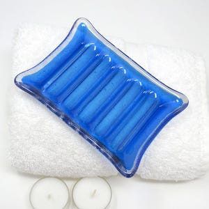 May include: A vibrant blue glass soap dish with a textured design, resting on a fluffy white towel. The soap dish has a unique shape and is surrounded by two small white candles, creating a spa-like ambiance.
