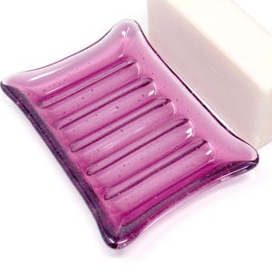 May include: A vibrant, fuchsia-colored glass soap dish with a rectangular shape and raised ridges. A white bar of soap rests beside the dish. The soap dish is a decorative bathroom accessory.