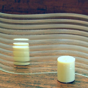 May include: A clear glass candle holder with a wavy design and etched lines. Two white candles are placed inside the holder.