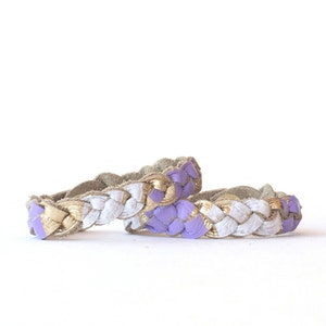 May include: Two braided leather bracelets in a white and purple colour scheme. The bracelets have gold accents.