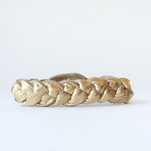 May include: A gold braided leather bracelet. The bracelet is made of a thick, braided leather cord that is gold in colour.