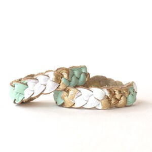May include: Two braided leather bracelets in mint green, white, and gold. The bracelets are stacked on top of each other.