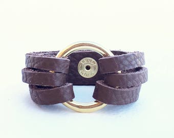 May include: A brown leather bracelet with a gold metal ring. The ring has a small metal disc in the center with the word "HAND" written on it.