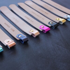 May include: A set of ten leather bookmarks with different colored metal tips. The bookmarks are all beige and have different text on them, including "BEST GRANOLA", "HAVE A NICE", and "TO GET CONNECTED".