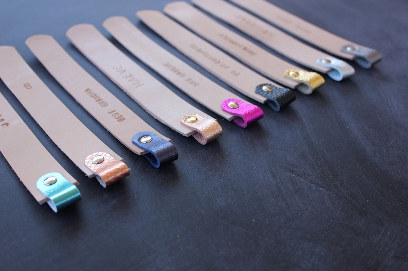 May include: A set of ten leather bookmarks with different colored metal tips. The bookmarks are all beige and have different text on them, including "BEST GRANOLA", "HAVE A NICE", and "TO GET CONNECTED".