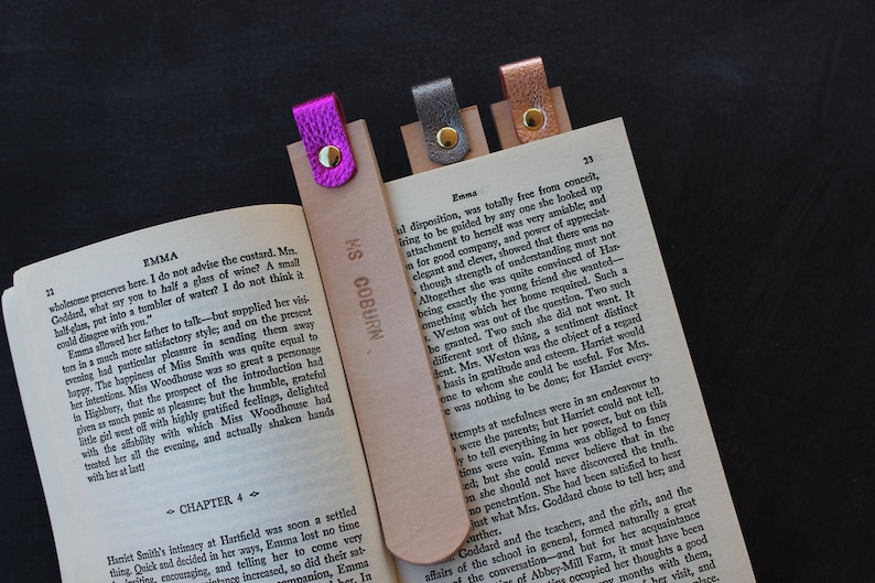 May include: Three leather bookmarks with metal accents. The bookmarks are pink, silver, and copper. The bookmarks are on a page of a book with the text "Emma" and "MS COBURN" printed on them.