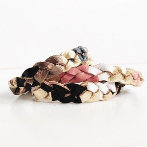 Può includere: Tre braccialetti in pelle intrecciati con dettagli dorati, neri, bianchi e rosa. I braccialetti sono impilati uno sopra l'altro.