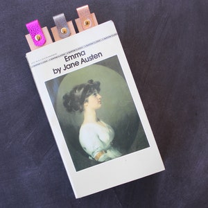May include: A paperback copy of the book "Emma" by Jane Austen with four colorful leather bookmarks. The bookmarks are pink, silver, gold, and brown.