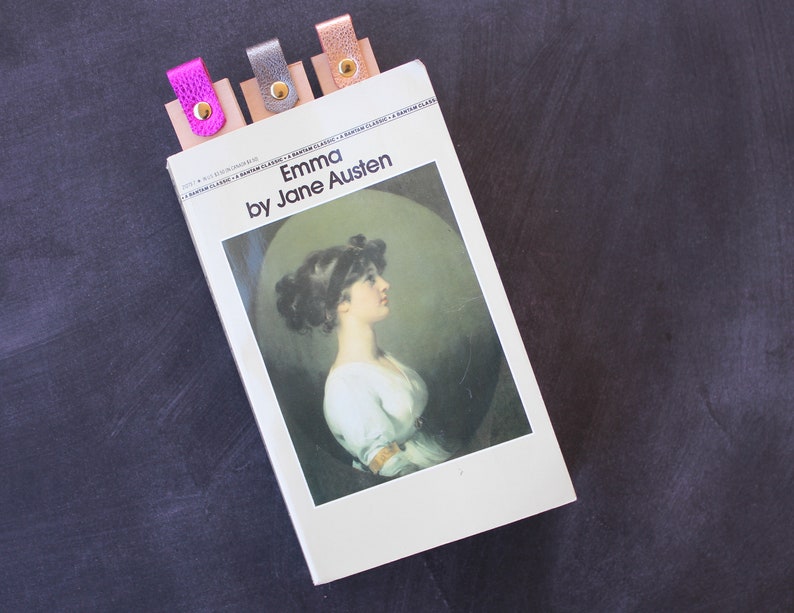 May include: A paperback copy of the book "Emma" by Jane Austen with four colorful leather bookmarks. The bookmarks are pink, silver, gold, and brown.