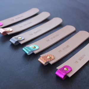 May include: Six leather bookmarks with different colored metal tips and personalized text. The bookmarks are beige with brown text. The metal tips are pink, gold, silver, teal, copper, and gold.