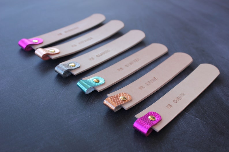 May include: Six leather bookmarks with different colored metal tips and personalized text. The bookmarks are beige with brown text. The metal tips are pink, gold, silver, teal, copper, and gold.
