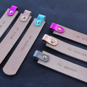 May include: Six leather bookmarks with different colored leather tabs and gold rivets. The bookmarks are labeled with names: "MS. COBURN", "HR. KROME", "MS. GRIMINGER", "MS. FREBOSA", "MS. MIRANDA", and "MS. CLAUDIO".