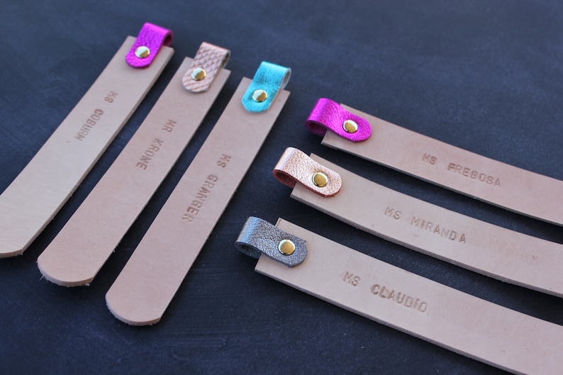May include: Six leather bookmarks with different colored leather tabs and gold rivets. The bookmarks are labeled with names: "MS. COBURN", "HR. KROME", "MS. GRIMINGER", "MS. FREBOSA", "MS. MIRANDA", and "MS. CLAUDIO".