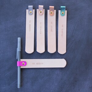 May include: Five leather bookmarks with different colored accents and names printed on them. The bookmarks are arranged in a row on a dark gray surface. A pen with a pink accent is attached to a sixth bookmark.