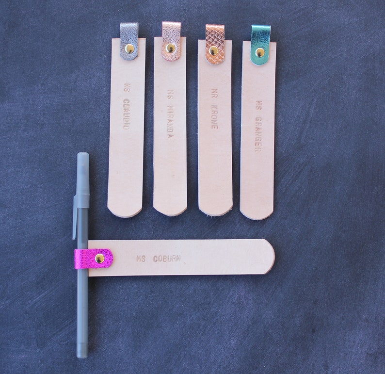 May include: Five leather bookmarks with different colored accents and names printed on them. The bookmarks are arranged in a row on a dark gray surface. A pen with a pink accent is attached to a sixth bookmark.