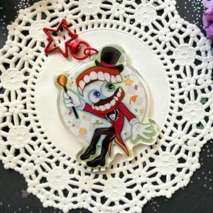 Puede incluir: Un charm de acrílico transparente con un personaje de dibujos animados con una boca grande, un ojo y un sombrero de copa. El personaje viste un traje rojo y sostiene una varita. Un cierre rojo en forma de estrella está unido al charm.