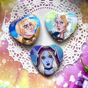 Puede incluir: Tres pines en forma de corazón con ilustraciones de personajes de fantasía. El primer pin muestra a un hombre rubio con ojos azules que lleva una armadura. El segundo pin muestra a una mujer rubia con ojos azules que lleva un atuendo blanco y dorado. El tercer pin muestra a una mujer con el pelo largo morado y ojos azules que lleva un atuendo morado y azul.