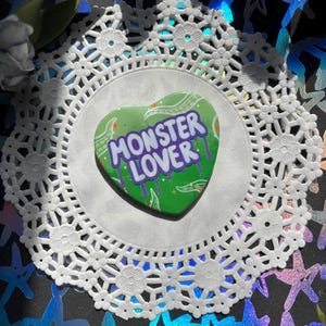 May include: Green heart-shaped pin with the text "Monster Lover" in purple and white letters with a dripping paint effect. The pin is on a white doily.