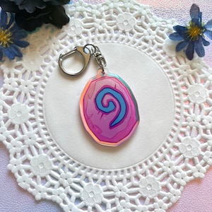 以下が含まれることがあります： 銀色のキーリングが付いたピンクとブルーの渦巻きキーホルダー。キーホルダーは丸みを帯びた楕円形で、ホログラフィック仕上げになっています。