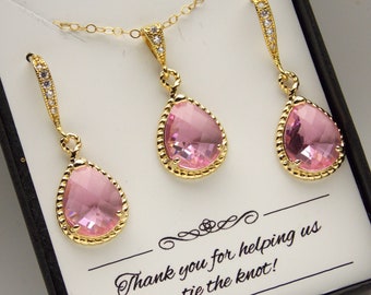 Puede incluir: Un conjunto de pendientes y un collar de color rosa en forma de lágrima con engastes dorados. Las joyas están en una caja negra con una tarjeta blanca que dice "¡Gracias por ayudarnos a atar el nudo!"