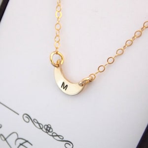 May include: A gold necklace with a crescent moon pendant. The pendant has the letter "M" engraved on it.