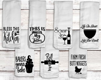 May include: White kitchen towels with black text and graphics. Designs include phrases like "Bless This Kitchen," "This Is My Jam," and "Farm Fresh Butt Nuggets." Other designs feature a wine bottle, rolling pin, and a cupcake.