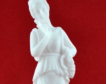 Puede incluir: Estatua de alabastro blanco de una mujer de pie sobre una base blanca. La mujer lleva una túnica fluida y sostiene un ramo de flores en su mano izquierda. La estatua está sobre un fondo rojo.