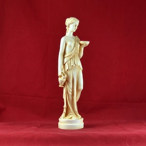 May include: A cream-coloured statue of a classical female figure holding a bowl and a jug. The statue is on a round base and is set against a red background. The figure is draped in flowing robes.