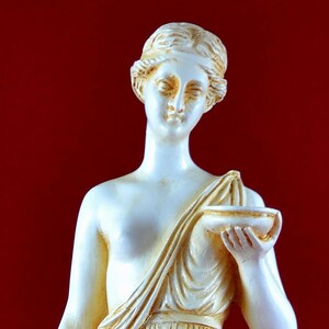 May include: A cream-coloured statue of a classical female figure, holding a small bowl and a vase. The statue is draped in flowing robes and stands on a white base against a red background.