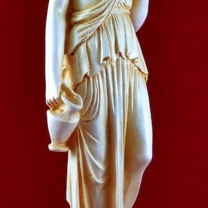 May include: A cream-coloured statue of a classical female figure holding a bowl and a vase. The statue is on a white base and is set against a red background. The figure is draped in flowing robes.