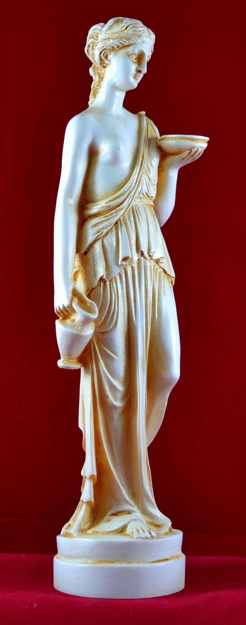 May include: A cream-coloured statue of a classical female figure holding a bowl and a vase. The statue is on a white base and is set against a red background. The figure is draped in flowing robes.