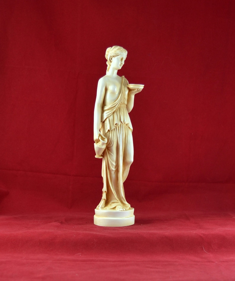 May include: A cream-coloured statue of a classical female figure holding a bowl and a jug. The statue is on a round base and is set against a red background. The figure is draped in flowing robes.