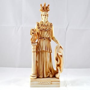 Puede incluir: Estatua de color crema de Atenea, la diosa griega de la sabiduría y la guerra. Lleva una corona y sostiene un escudo con una serpiente. Un búho se posa sobre una columna a su izquierda. La base de la estatua dice "ATHNA".