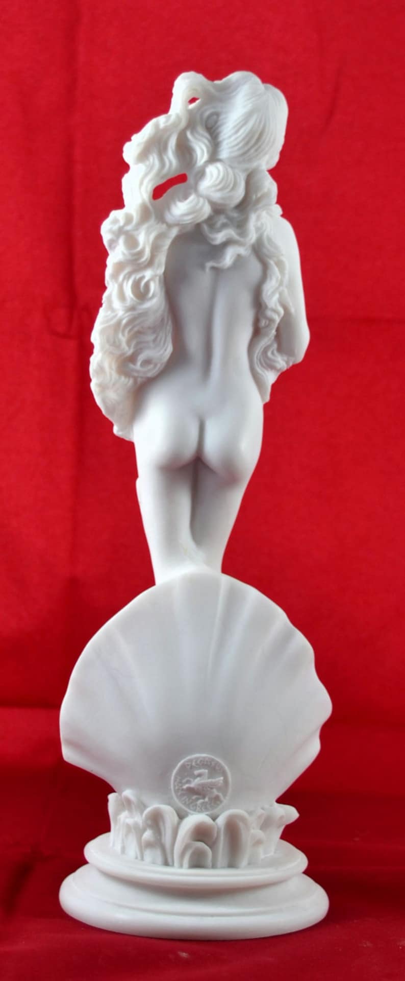 May include: White marble statue of a woman standing on a shell. The statue is a replica of the famous Venus de Milo sculpture. The statue is on a white pedestal with a circular base.