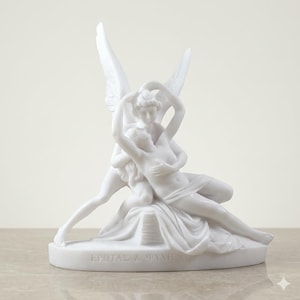 Puede incluir: Una escultura de mármol blanco que representa a Eros y Psique abrazándose. La escultura presenta dos figuras, una con grandes alas, entrelazadas en un abrazo amoroso. La base de la escultura tiene el texto "EΡΩΣ & ΨΥΧΗ".