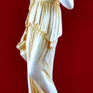May include: A cream-coloured statue of a classical female figure holding a small bowl. The statue has detailed drapery and is set against a red background. The figure is standing on a round base.