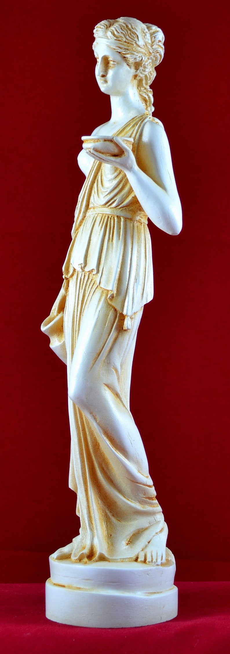May include: A cream-coloured statue of a classical female figure holding a small bowl. The statue has detailed drapery and is set against a red background. The figure is standing on a round base.