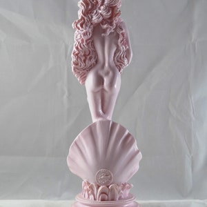 May include: A pink ceramic statue of the Venus de Milo standing on a shell base. The statue is a replica of the famous Greek sculpture.